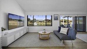A modern living room with a large TV, a round coffee table, a grey sofa, and a view of a beach and houses through large windows. - Deja Bleu - family home with private jetty (Port Macquarie)