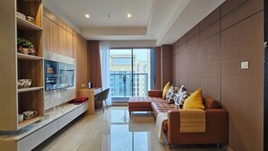 A modern living room with a sofa set, a TV mounted on a wooden cabinet, and a view of the city through large windows. - Branz Apartment BSD City by Nagisa Bali (Pagedangan)