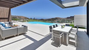 A modern outdoor patio with a pool, seating area, and a view of the coastline. - Villa in Salobre With Pool & Golf Nearby (San Bartolomé de Tirajana)