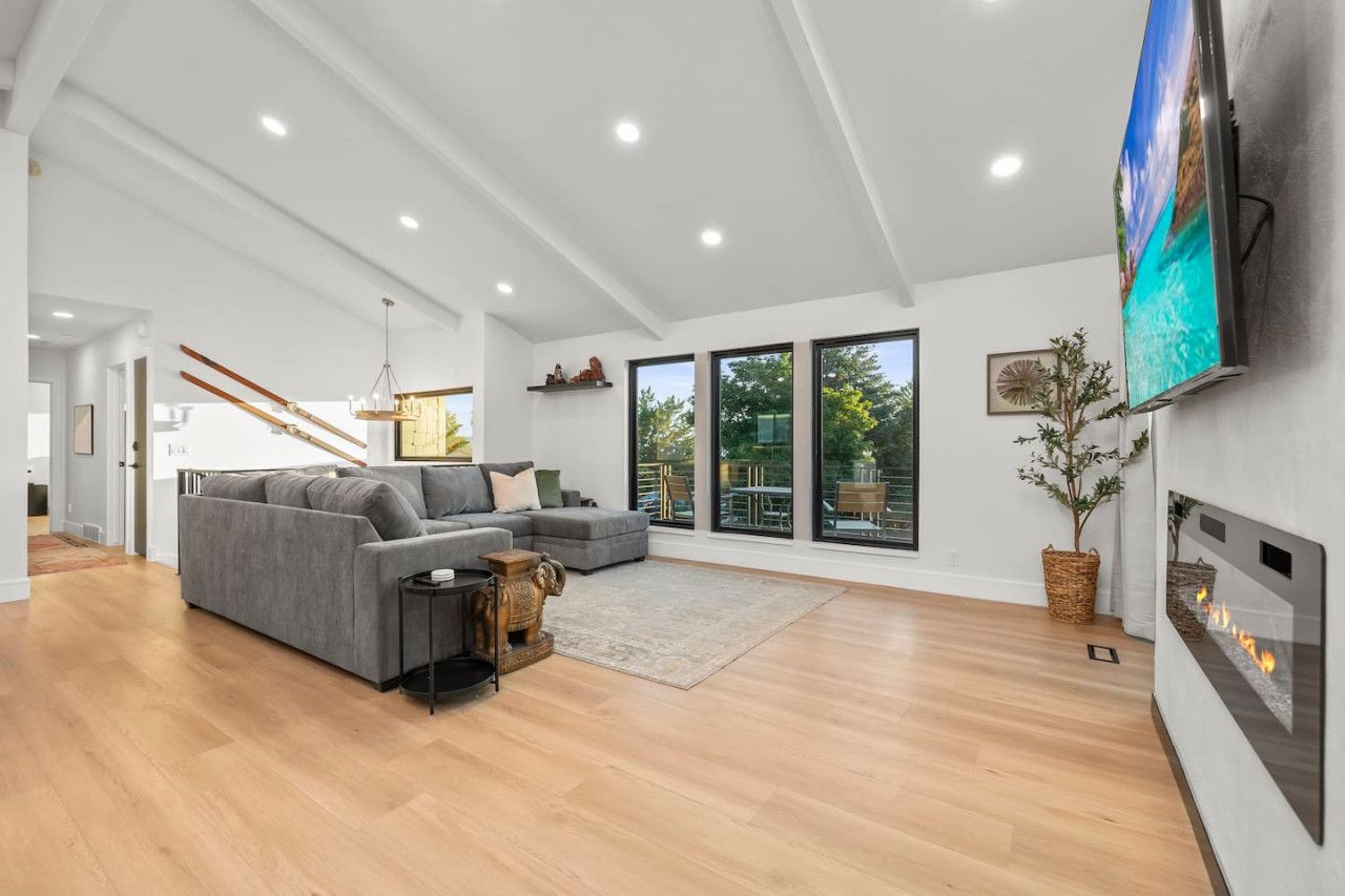 Un salon spacieux avec un canapé gris, un plancher en bois, une cheminée et de grandes fenêtres donnant sur l’extérieur.