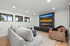 Una sala moderna con un sofá gris, un sofá marrón y un televisor de pantalla plana montado en una pared negra. Hay tres ventanas y una chimenea.