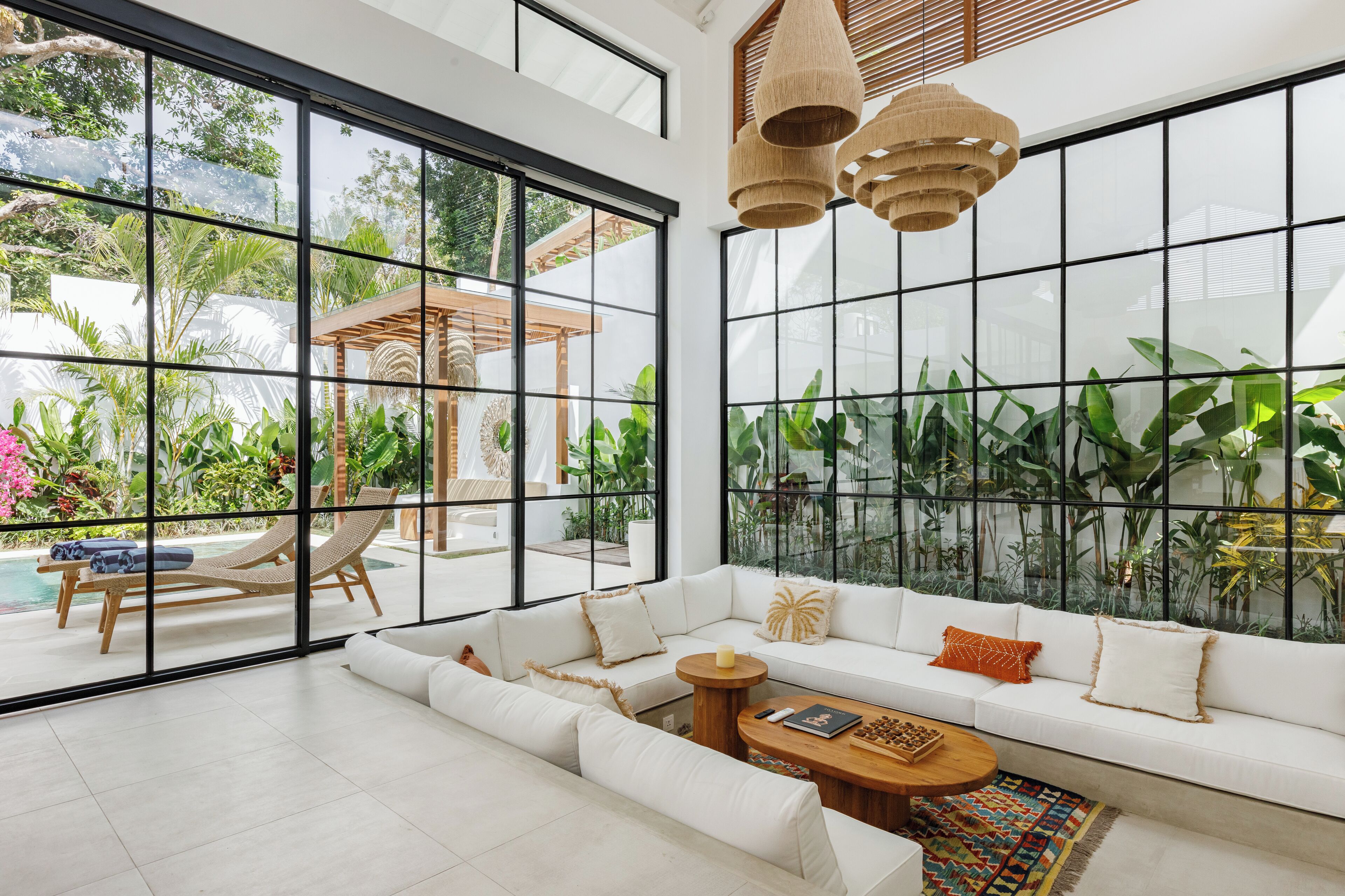 A spacious living area with a large white sofa, a wooden coffee table, and a view of a pool and lush greenery through floor-to-ceiling windows.