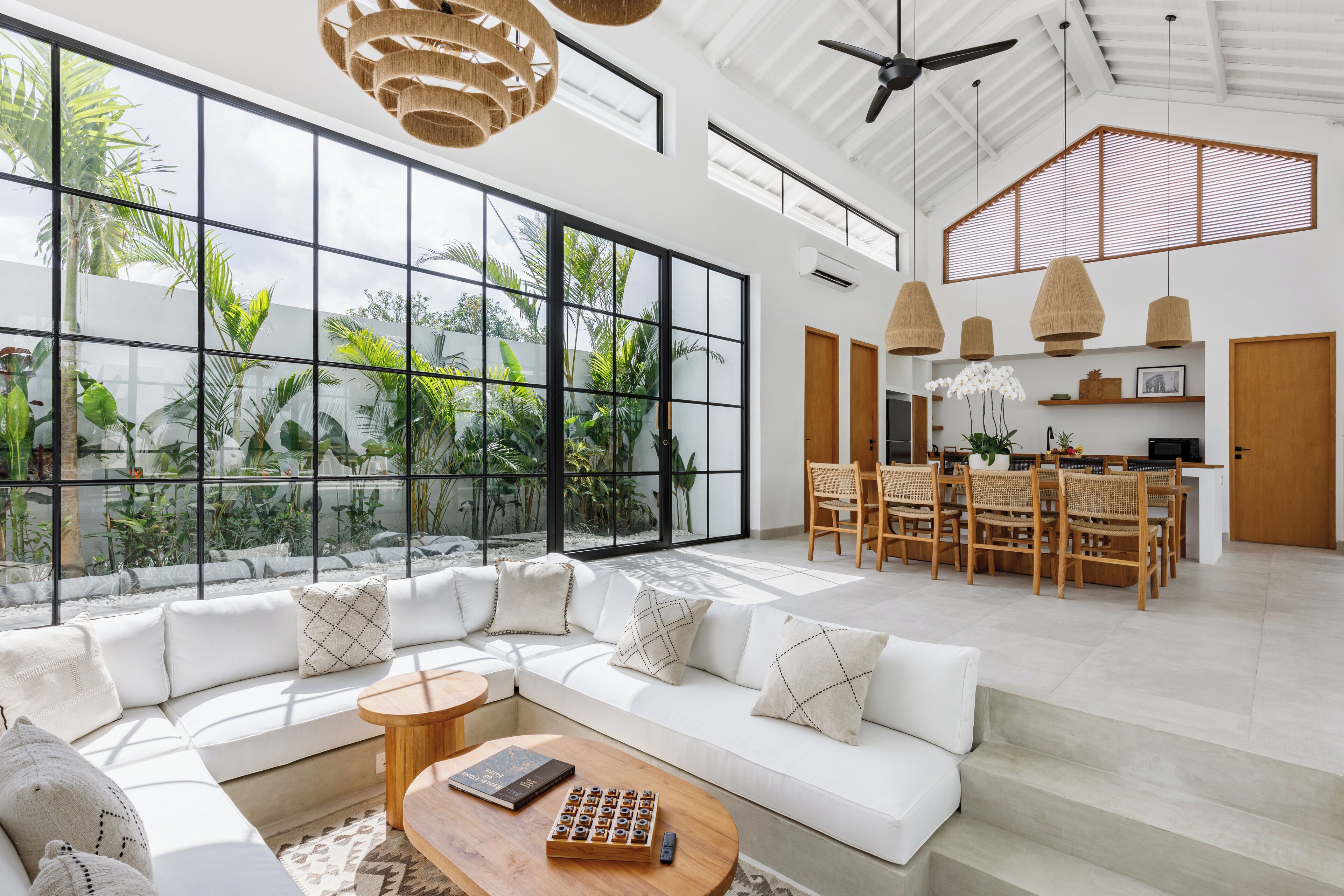 A spacious living area with a large window overlooking a tropical garden, a modern kitchen, and a dining area with wooden furniture.