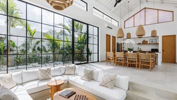 A spacious living area with a large window overlooking a tropical garden, a modern kitchen, and a dining area with wooden furniture.
