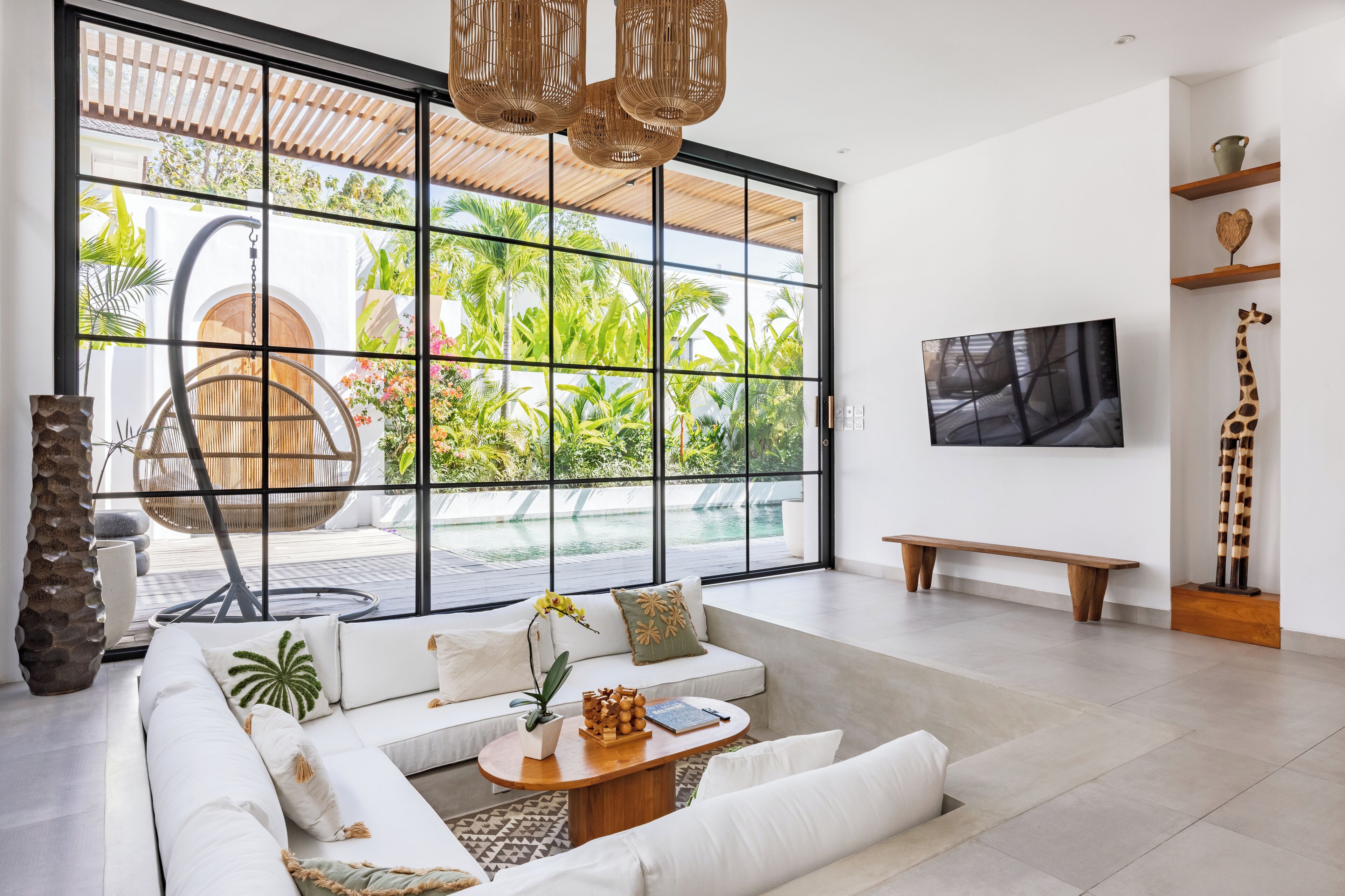 Un salón espacioso con un amplio ventanal corredero que da a una zona de piscina, un conjunto de sofás cómodos, una mesa de centro de madera y un televisor montado en la pared.
