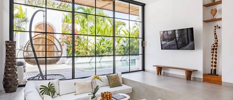 Un salón espacioso con un amplio ventanal corredero que da a una zona de piscina, un conjunto de sofás cómodos, una mesa de centro de madera y un televisor montado en la pared.