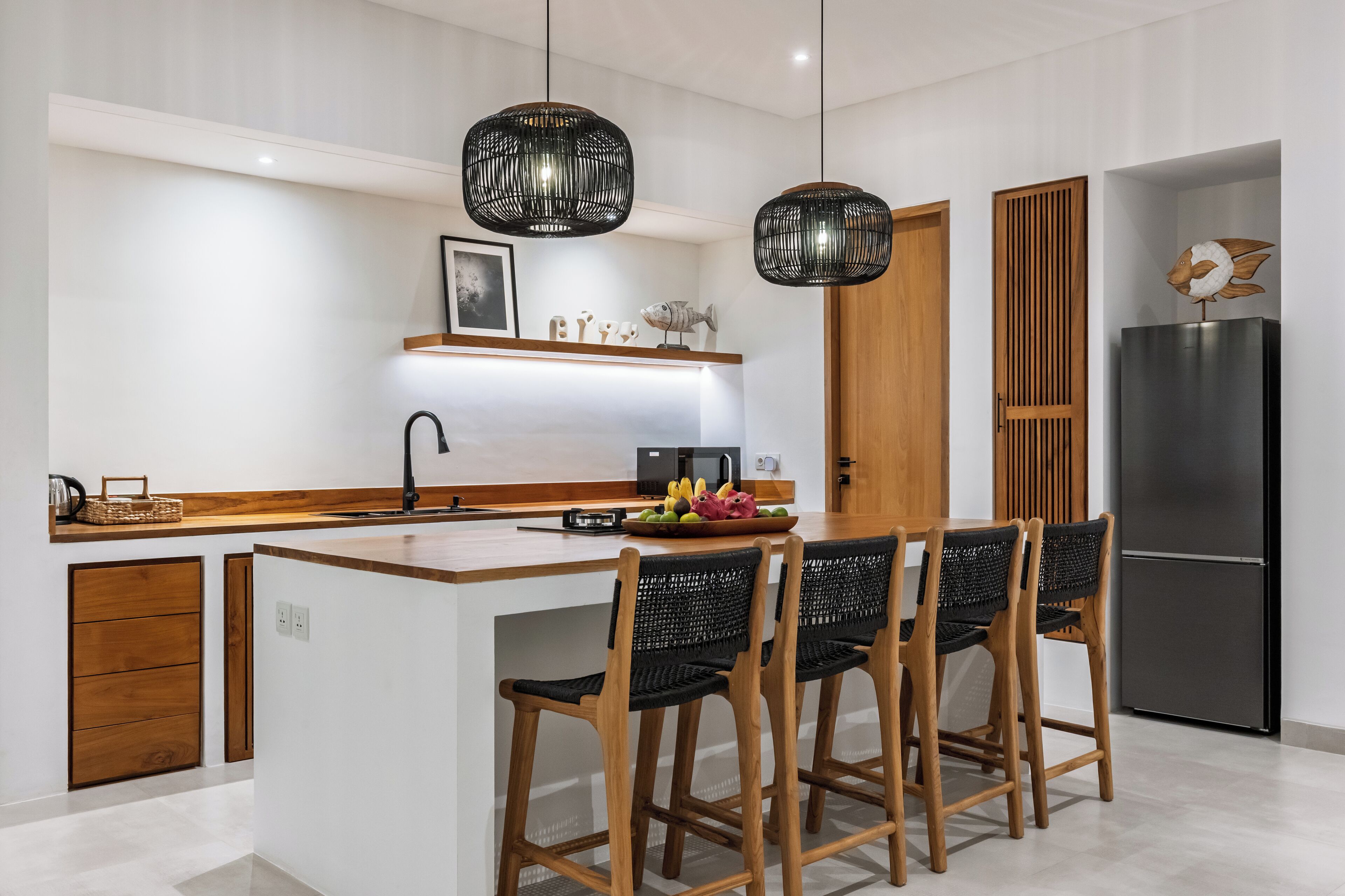 Une cuisine moderne dotée d’un îlot central, de luminaires suspendus noirs en forme de suspensions tissées et d’un réfrigérateur en acier inoxydable.