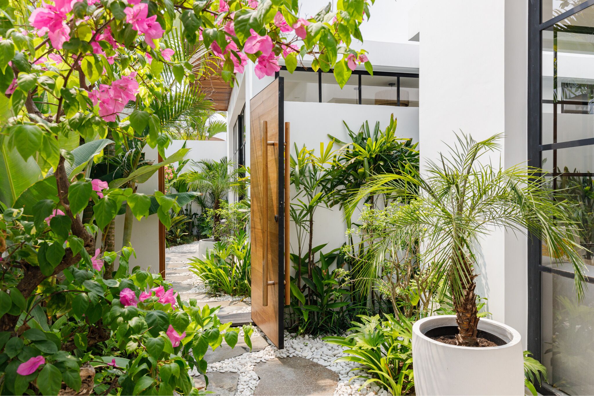 A modern building with a wooden door, surrounded by lush greenery and pink flowers.