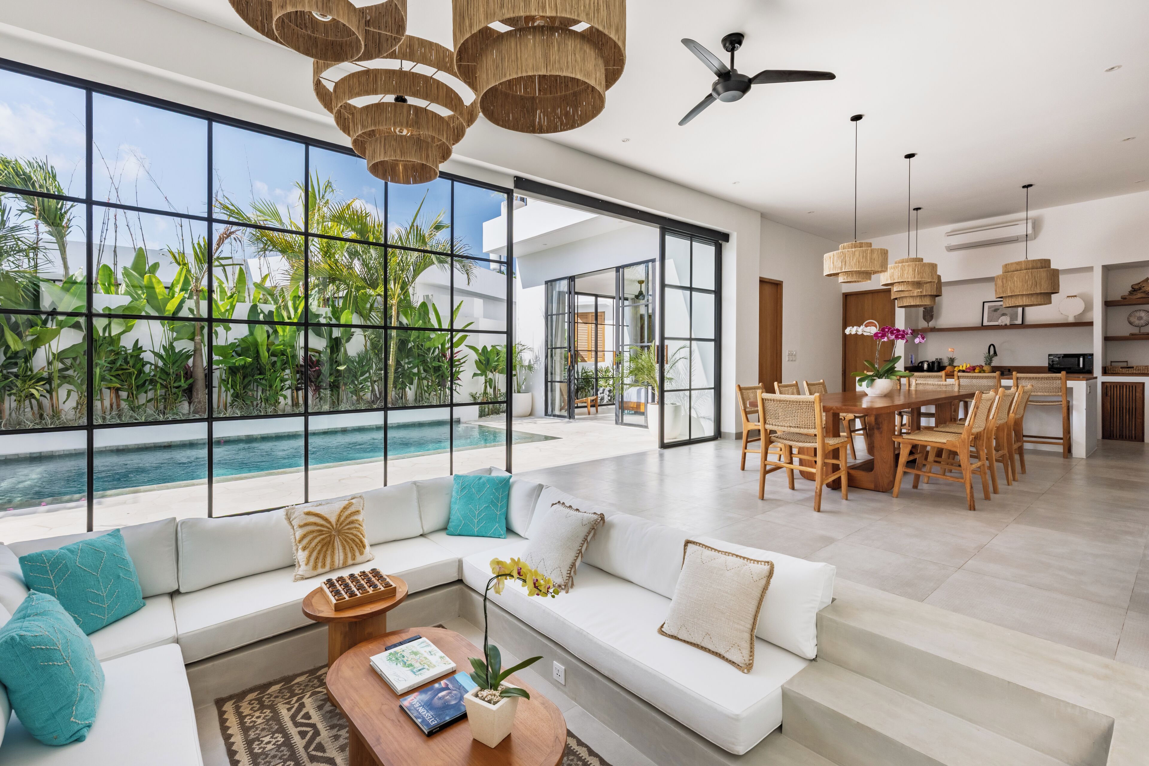 A modern living room with a large window overlooking a pool, a dining area with wooden furniture, and a ceiling fan.