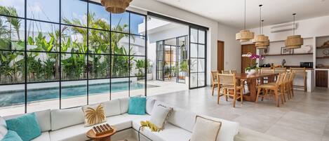 A modern living room with a large window overlooking a pool, a dining area with wooden furniture, and a ceiling fan.