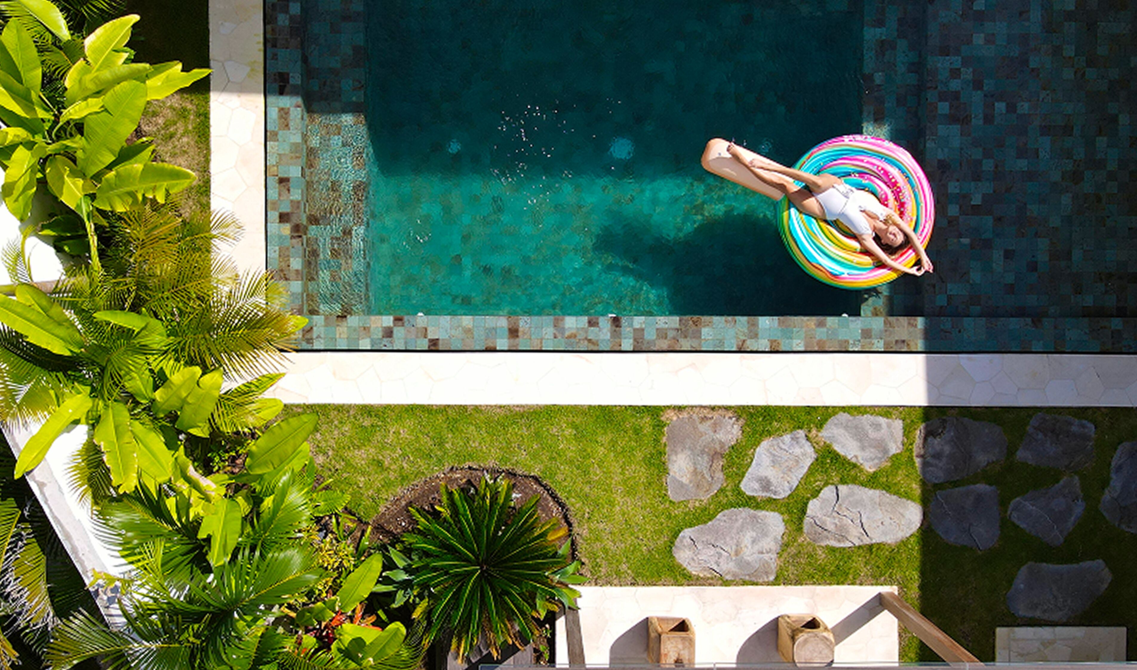 A swimming pool with a person floating on a colorful ring, surrounded by lush greenery and stone pathways.