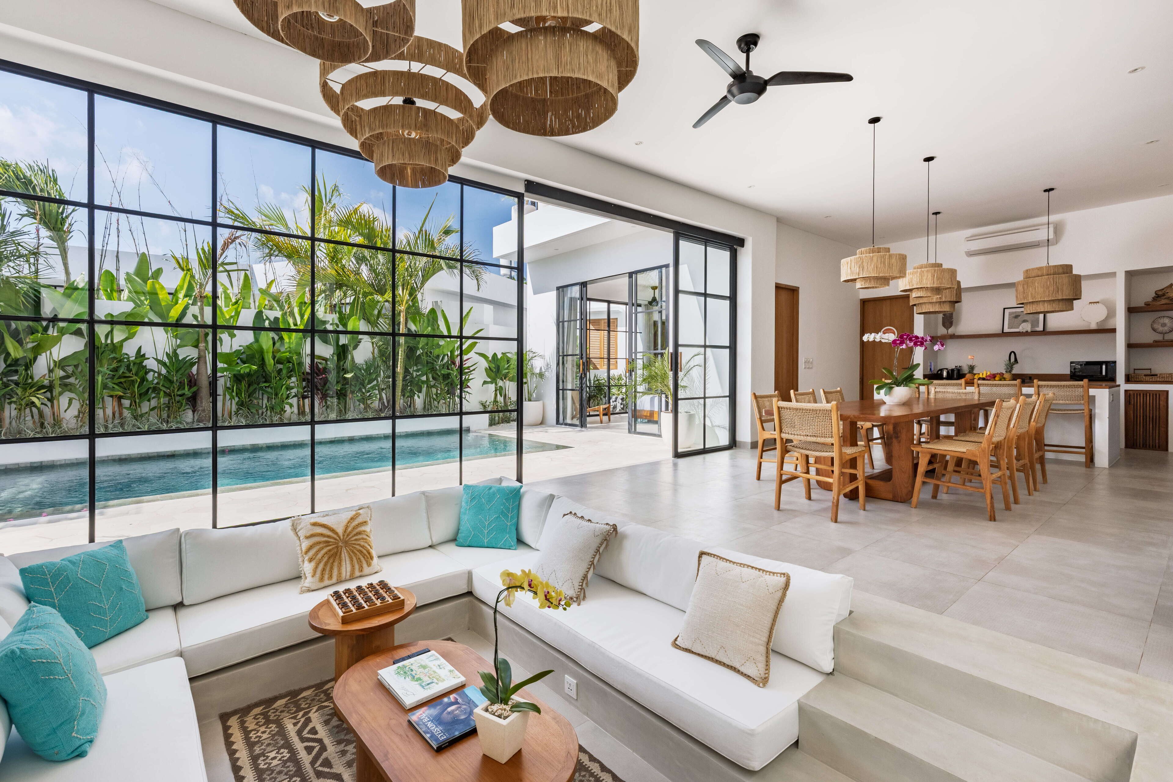 A modern living room with a large window overlooking a pool, a dining area with wooden furniture, and a ceiling fan.