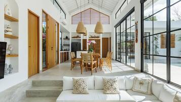 Ein geräumiger Wohnbereich mit weißem Sofa, Esstisch aus Holz und großen Fenstern mit Blick ins Freie.