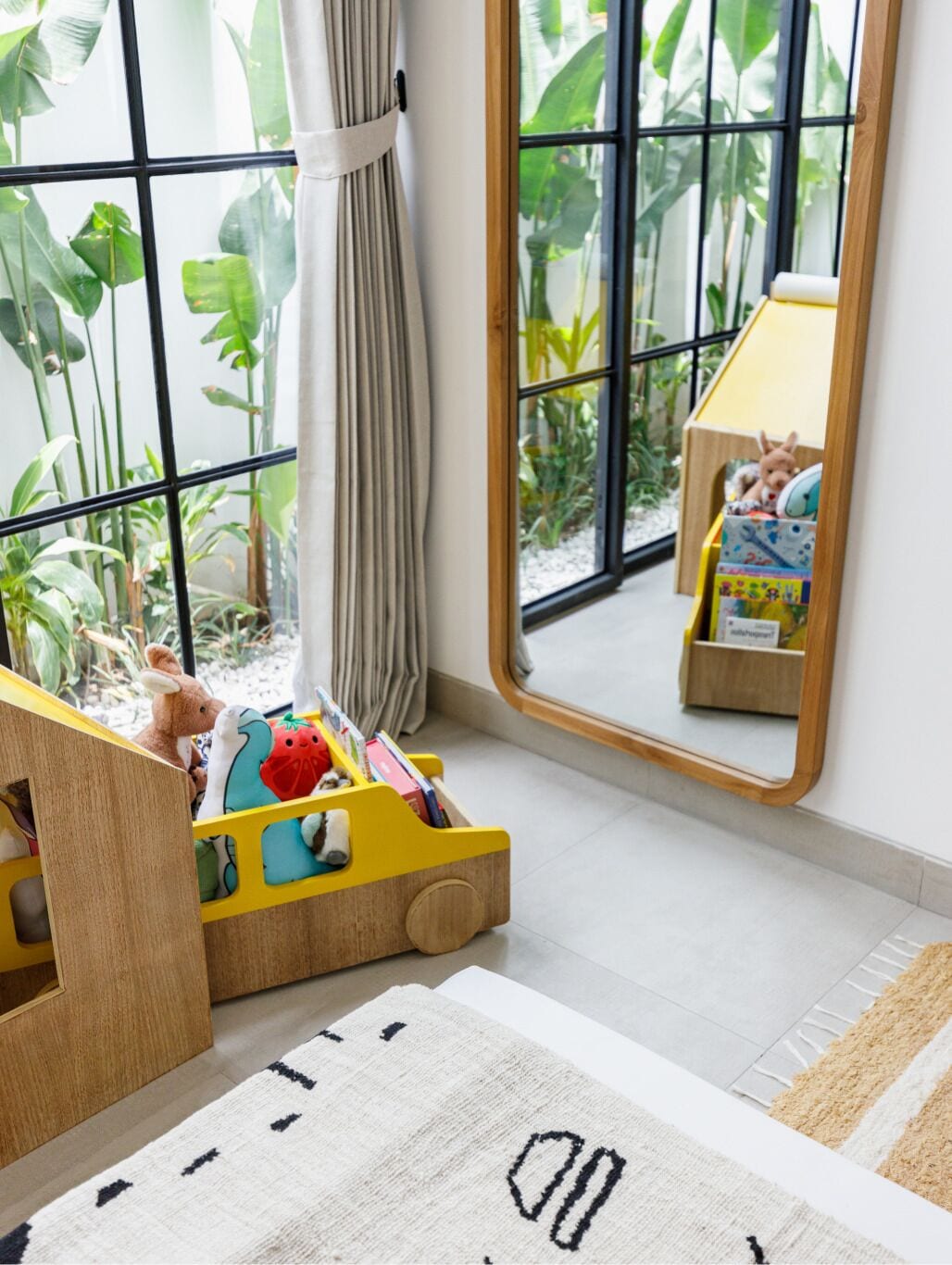 Zona infantil con un cofre de juguete de madera, un espejo que refleja la habitación y ventanas amplias con plantas tropicales.