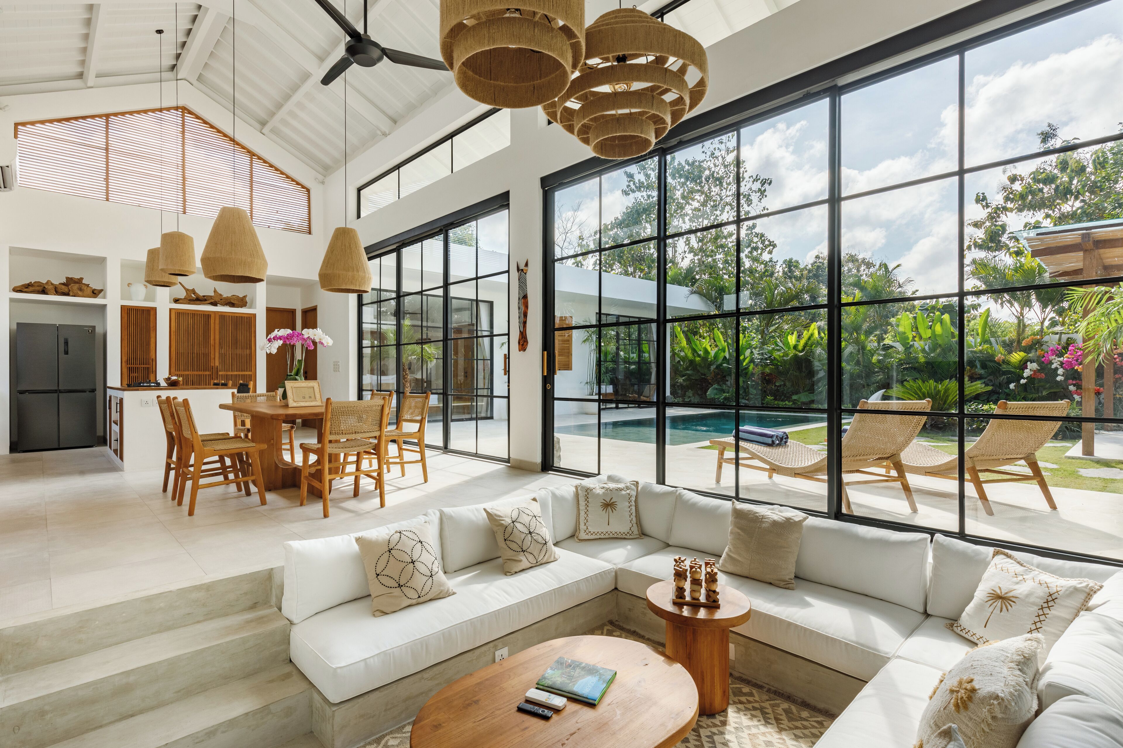 A spacious living area with a large window overlooking a pool and garden, a dining area with wooden furniture, and a modern kitchen.