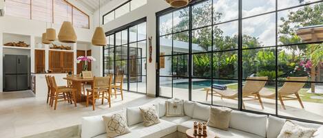 A spacious living area with a large window overlooking a pool and garden, a dining area with wooden furniture, and a modern kitchen.