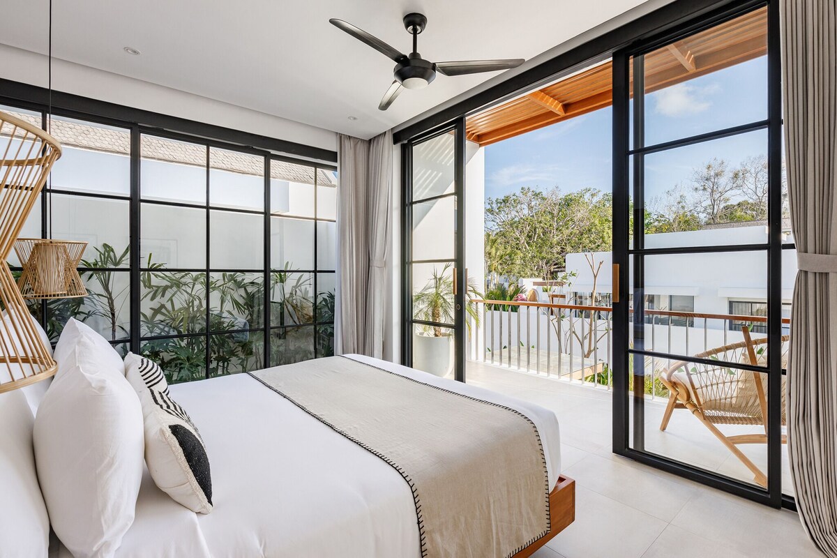 A modern bedroom with a large bed, a ceiling fan, and a balcony with a view of greenery and a building.