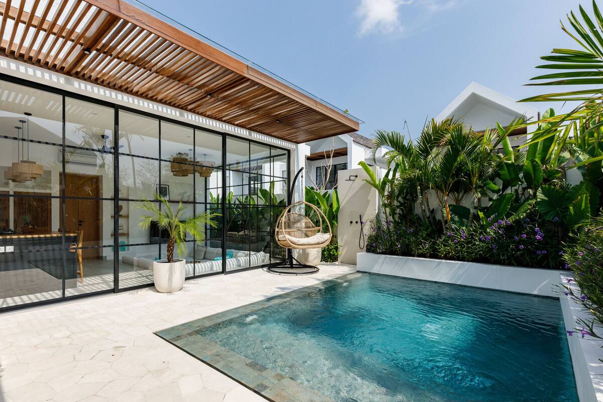 A modern house with a glass-enclosed patio, a pool, and lush greenery.