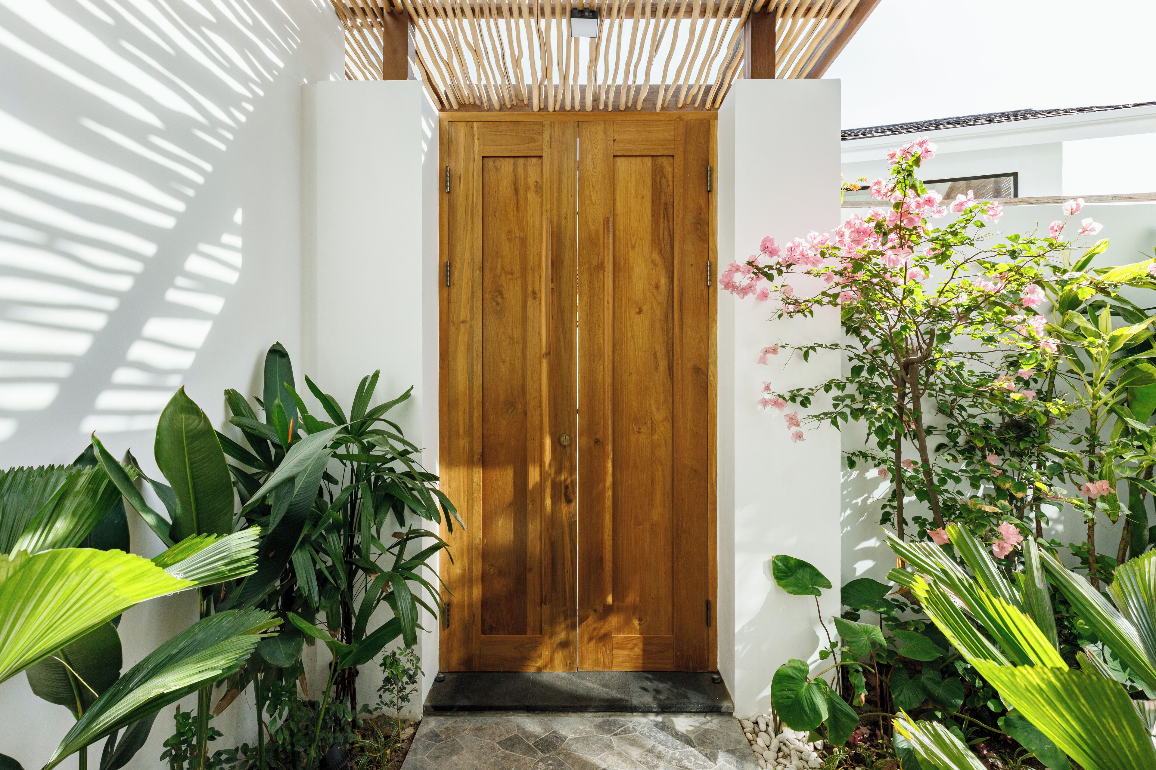 A wooden door with a stone step leading up to it, surrounded by lush green plants and a pink flowering tree.