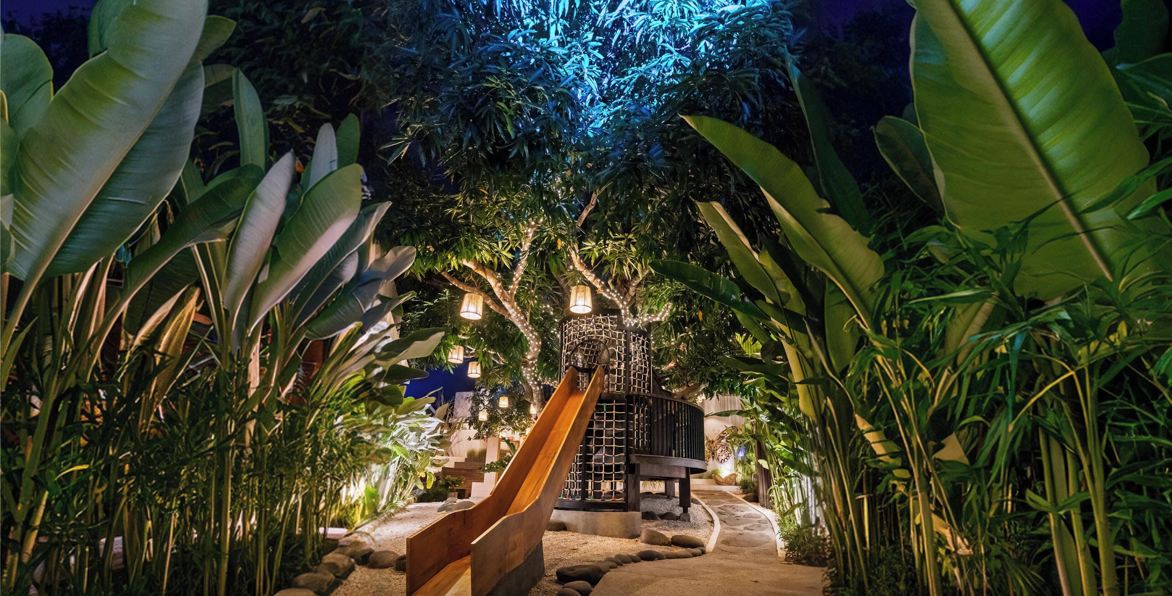 Ein Garten bei Nacht mit einer Holzrutsche, umgeben von üppigen tropischen Pflanzen und beleuchtet mit Lichterketten.