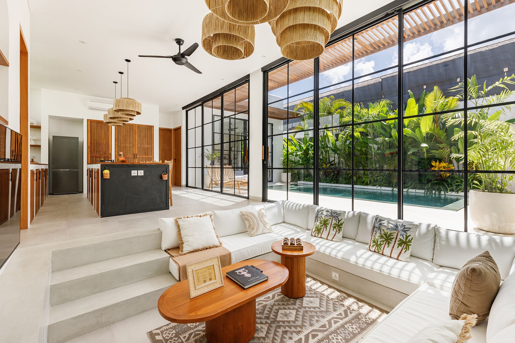 A modern living room with a large window, a sofa set, a coffee table, and a kitchen area visible through the glass.