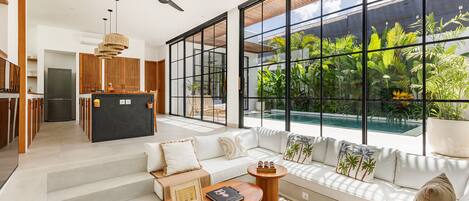 A modern living room with a large window, a sofa set, a coffee table, and a kitchen area visible through the glass.