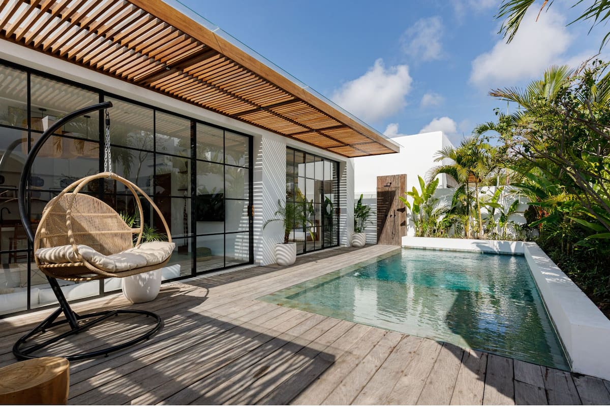A modern outdoor pool area with a wooden deck, a wicker chair, and a glass-enclosed building.
