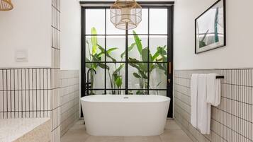 A modern bathroom with a freestanding bathtub, a large window with plants, and a hanging light fixture.