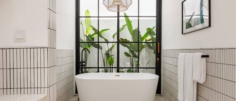 Une salle de bain moderne avec une baignoire sur pieds, une grande fenêtre agrémentée de plantes et un luminaire suspendu.