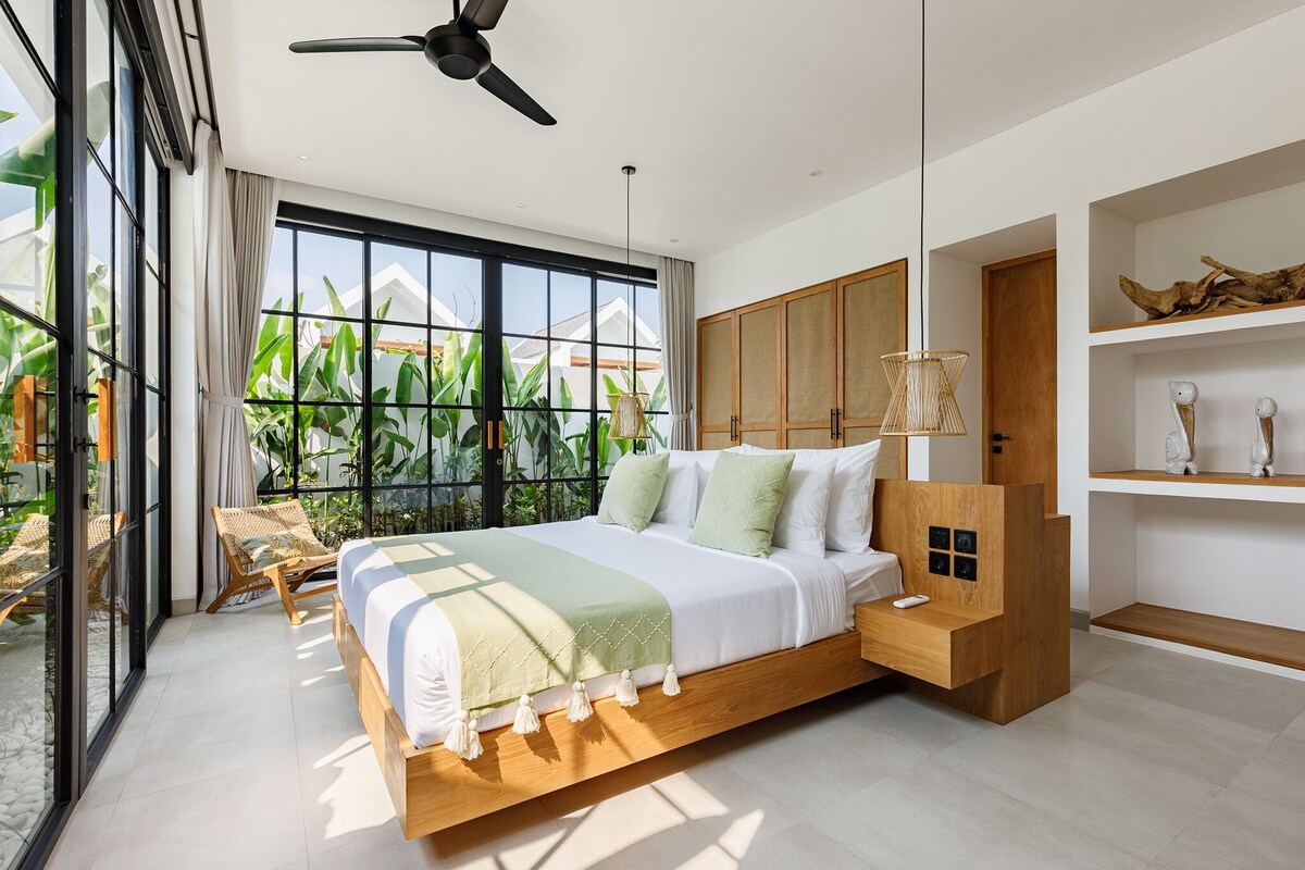 A modern bedroom with a large bed, wooden headboard, and a view of greenery outside.