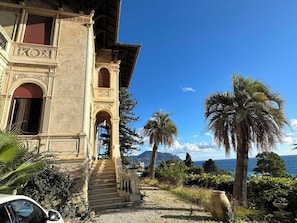 Un edificio storico con una maestosa scalinata che conduce a un ingresso decorato, circondato da rigogliosa vegetazione e vista sul mare.