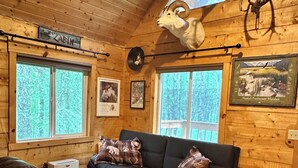 A cabin interior with a black leather sofa, a leather chair, wooden walls, and a mounted animal head. - Rustic and cozy cabin with all new amenities near beautiful Hatcher’s Pass (Palmer)