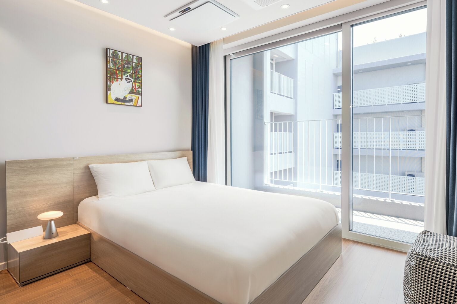 A modern hotel room with a large bed, a bedside table with a lamp, a window with a view of the building, and a painting on the wall.