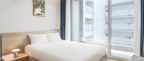 A modern hotel room with a large bed, a bedside table with a lamp, a window with a view of the building, and a painting on the wall.