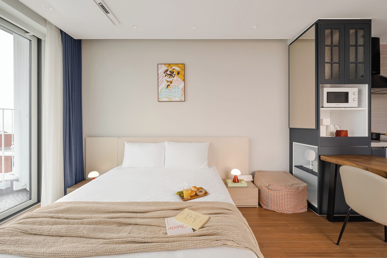 A modern hotel room with a large bed, bedside tables, a desk, and a kitchenette.