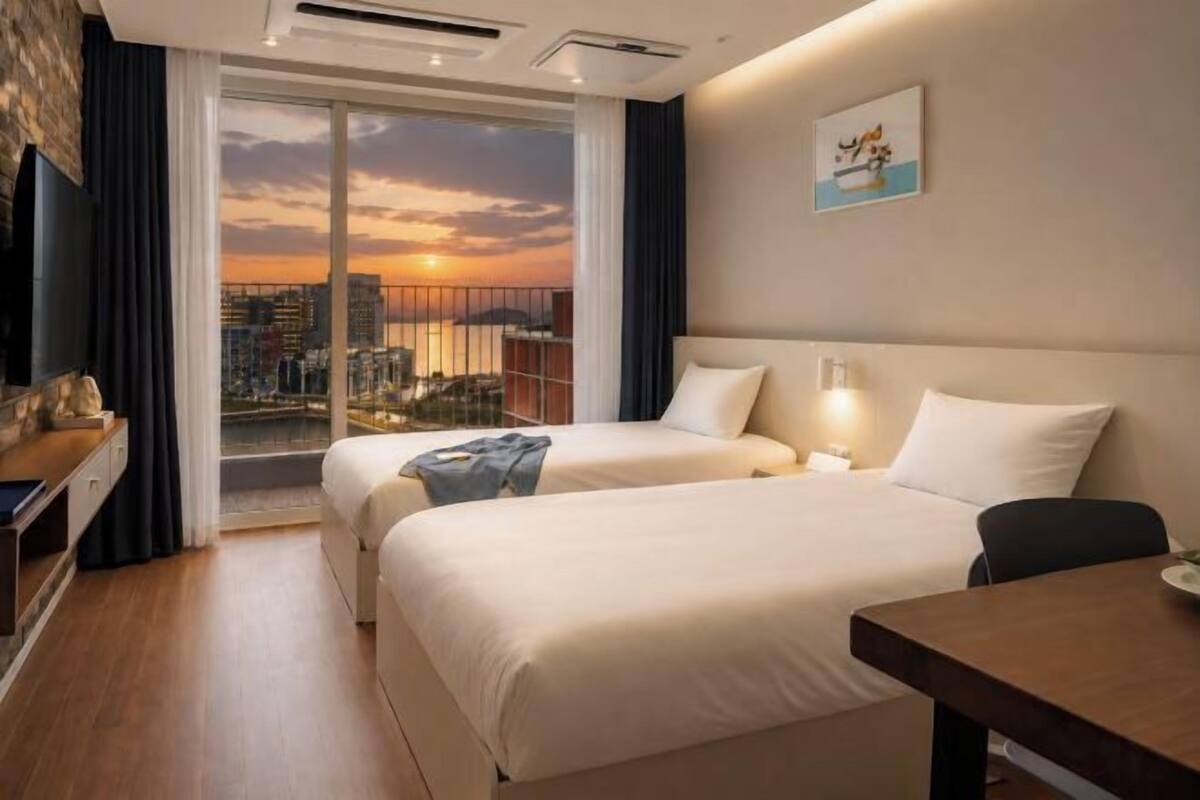 A hotel room with two beds, a desk, a chair, a TV, and a balcony with a city view.