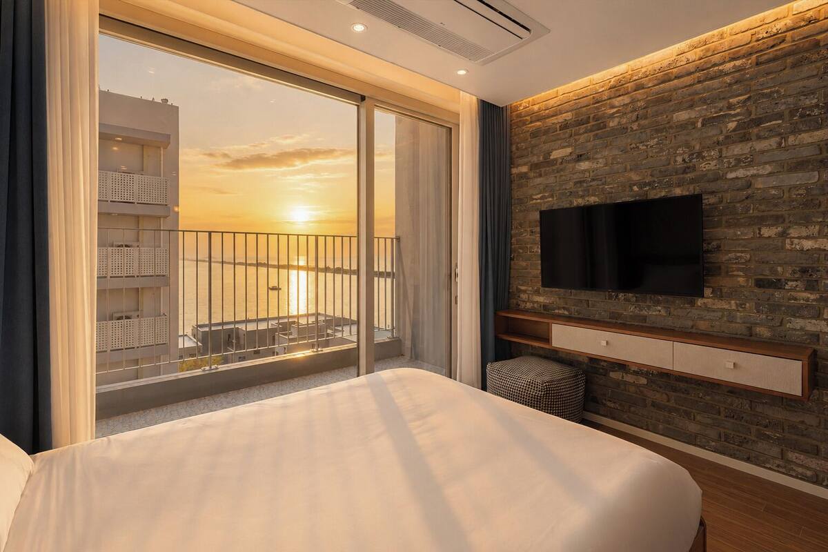 A hotel room with a large bed, a TV mounted on a brick wall, a balcony with a view of the sunset, and a sliding glass door.