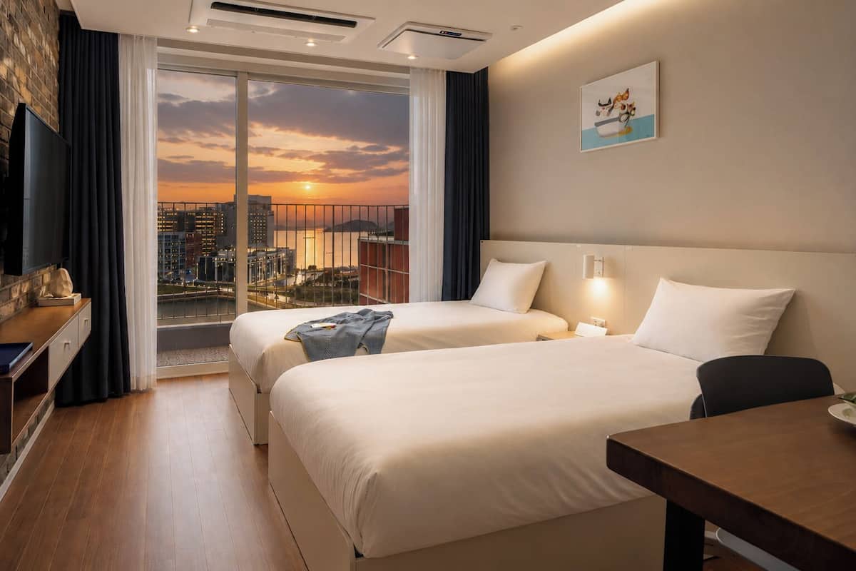 A hotel room with two beds, a desk, a TV, and a city view at sunset.