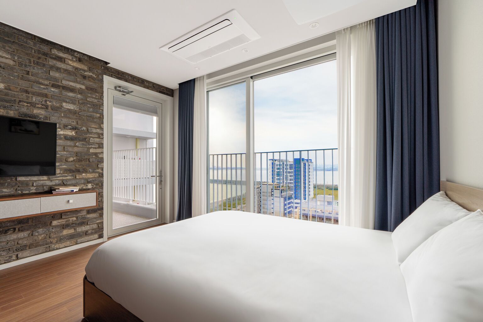 A hotel room with a large bed, a TV on a brick wall, a balcony with a view of buildings, and a window with curtains.