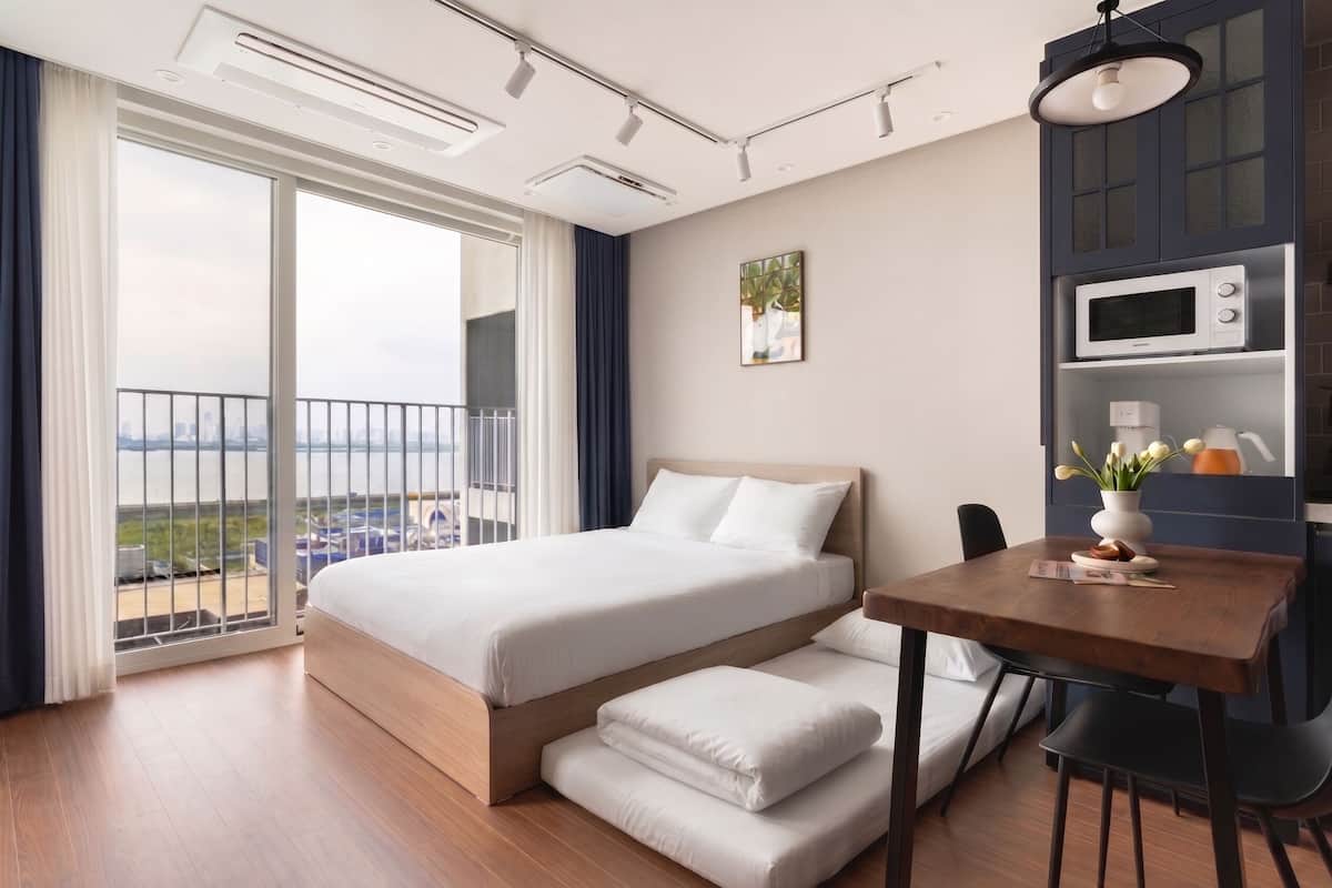 A modern hotel room with a large bed, a dining table, a microwave, and a view of the city.
