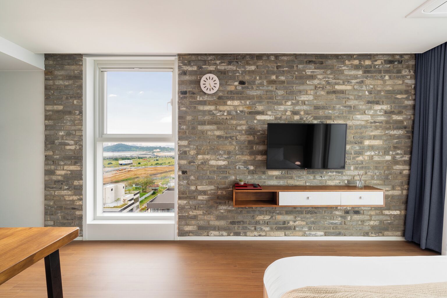 A modern bedroom with a brick wall, a mounted TV, a window with a view of a landscape, and a wooden bedside table.