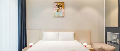 A modern hotel room with a large bed, bedside tables, a small seating area, and a painting on the wall.