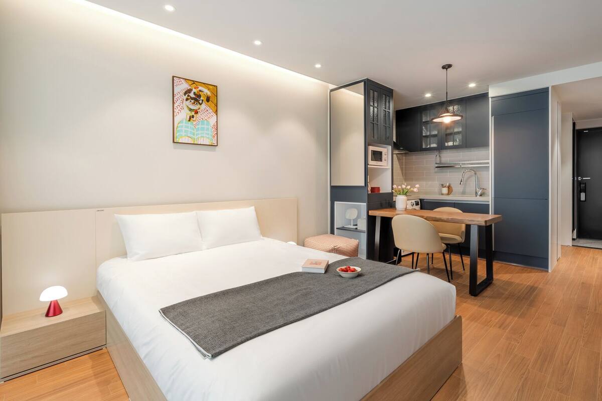 A modern hotel room with a large bed, a desk, and a kitchenette.