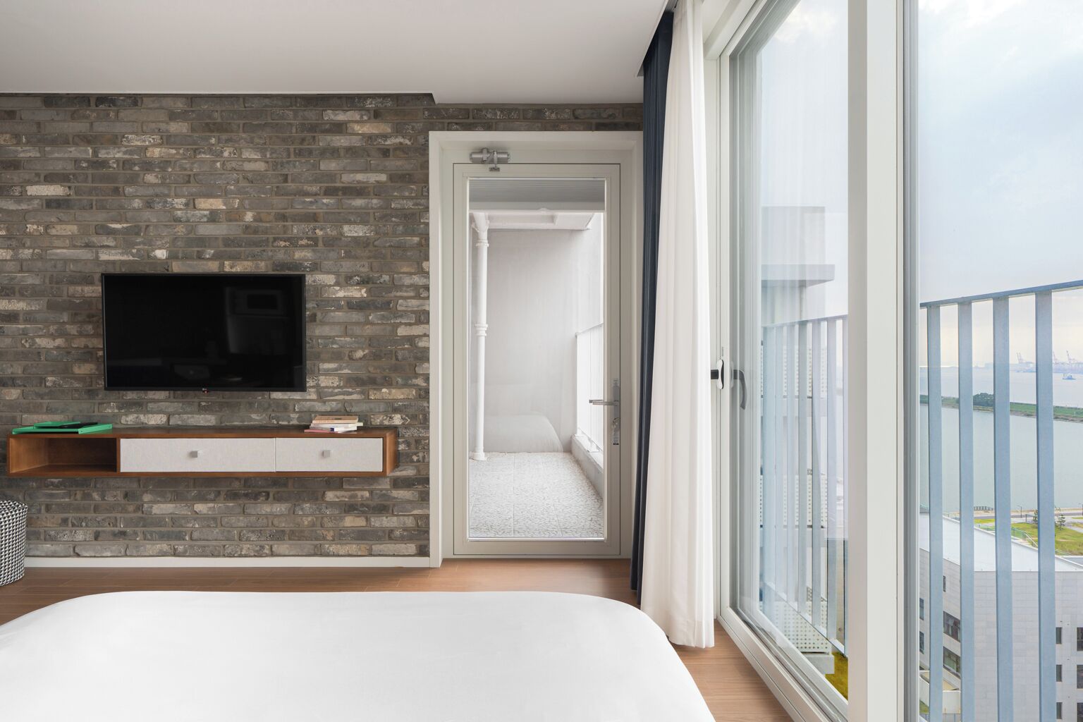 A modern bedroom with a brick wall, a flat-screen TV, a bed, and a balcony with a view.