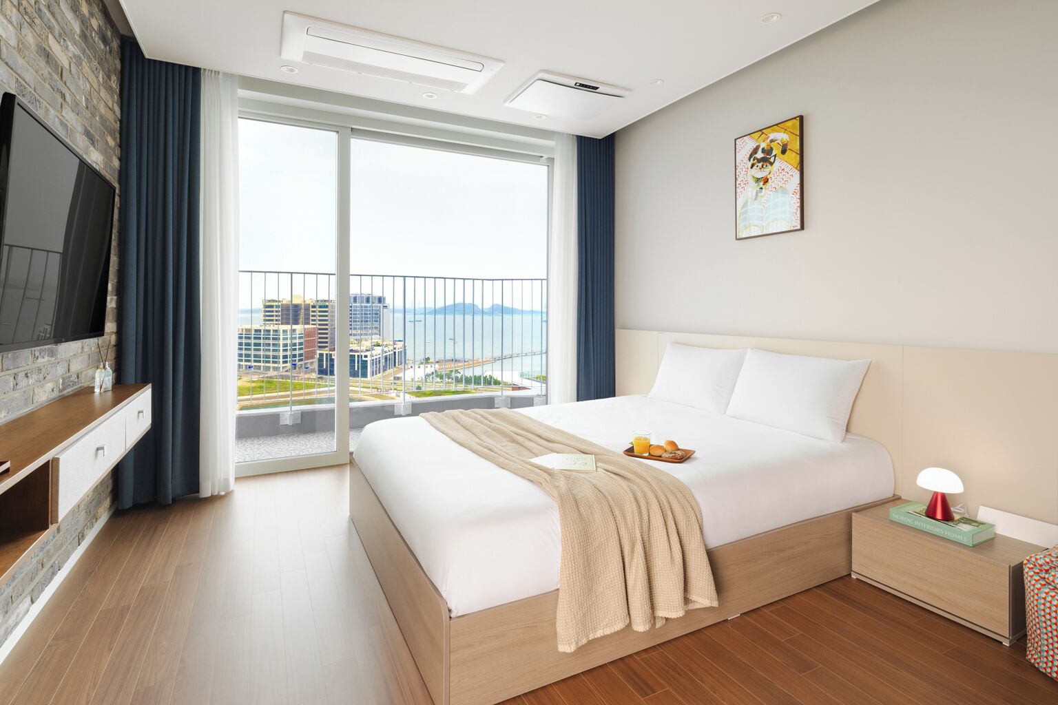Una moderna camera d'hotel con un grande letto, una TV fissata a una parete in mattoni, un balcone con vista sulla città e un comodino con una lampada.