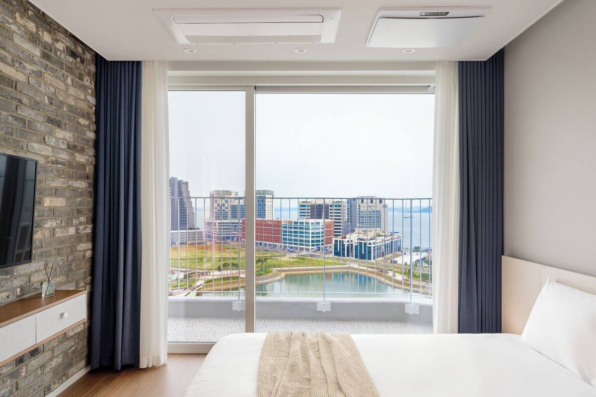 A modern hotel room with a large bed, a TV on the left wall, and a balcony with a city view.
