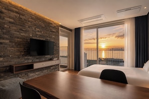 Camera d'hotel con parete in mattoni, televisore a schermo piatto, scrivania in legno, un letto e un balcone con vista sul tramonto.