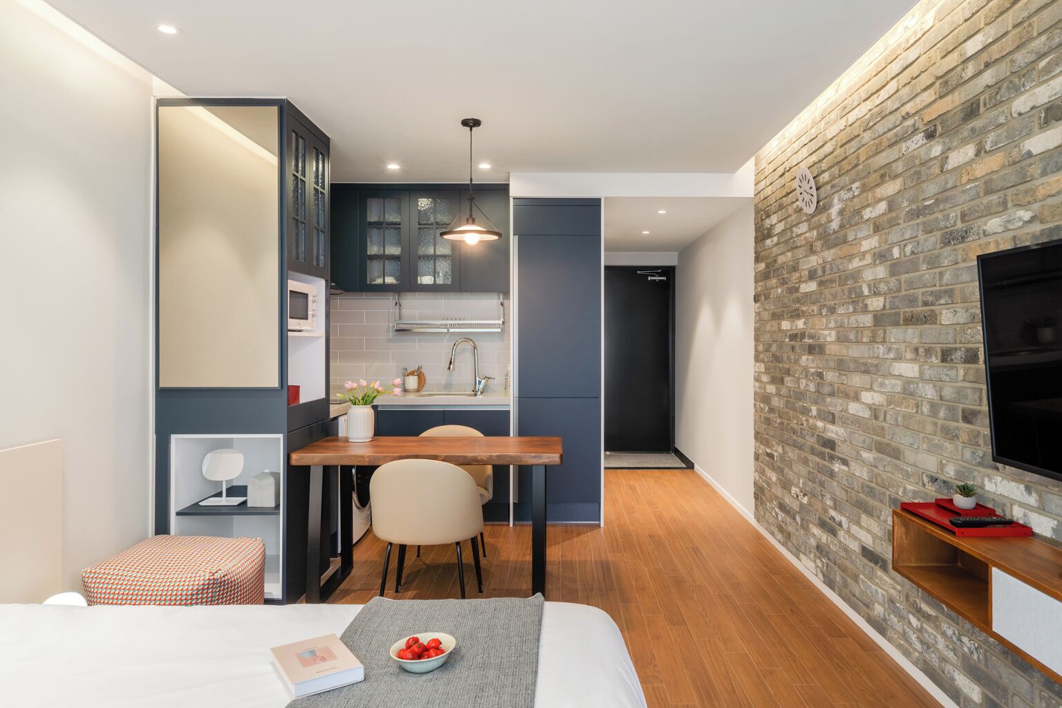 Una camera d'albergo compatta con angolo cottura, zona pranzo, un letto e una TV montata su un muro in mattoni.