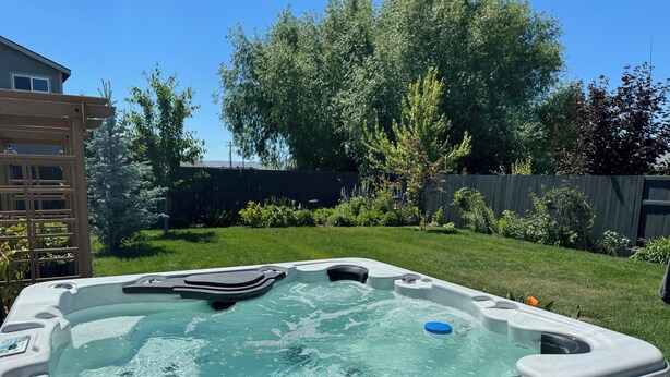 A hot tub in a backyard with a wooden deck, surrounded by a wooden fence, trees, and a grassy lawn.