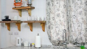 A kitchen with wooden shelves holding cups and a sign, a kettle, a faucet, and a patterned curtain. - Secret Place  (Gros islet)